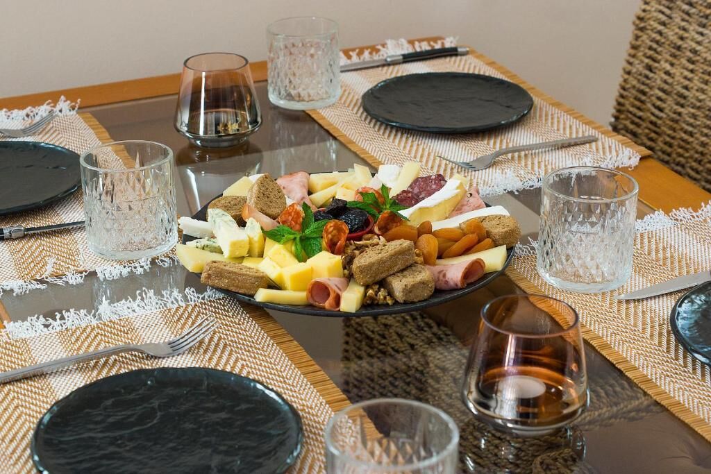A dining table with a cheese platter, glasses, cutlery, and a woven placemat.