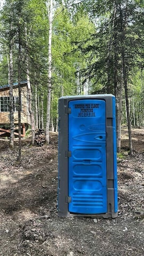 Eine mobile Toilette mit blauer Tür, beschriftet mit 'Around the Clock Pumping', in einem bewaldeten Gebiet.