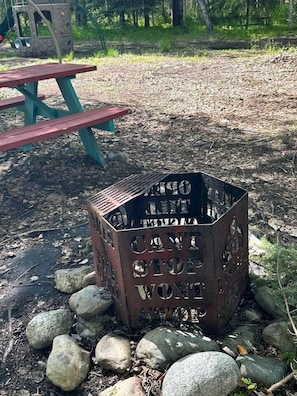 Ein metallenes Lagerfeuer mit der Aufschrift 'CAMPING CAN'T STOP WONDER', umgeben von Steinen und einem Picknicktisch in einer bewaldeten Gegend.