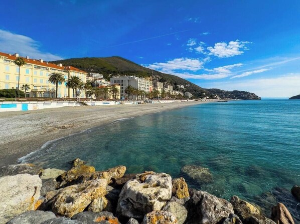 Une plage cĂŽtiĂšre avec une eau claire, un premier plan rocheux et des bĂątiments le long de la cĂŽte.