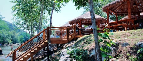 A wooden staircase leading to a thatched-roof structure by a river, surrounded by trees.