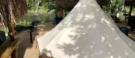 A large tent with a peaked roof, surrounded by trees and a body of water.