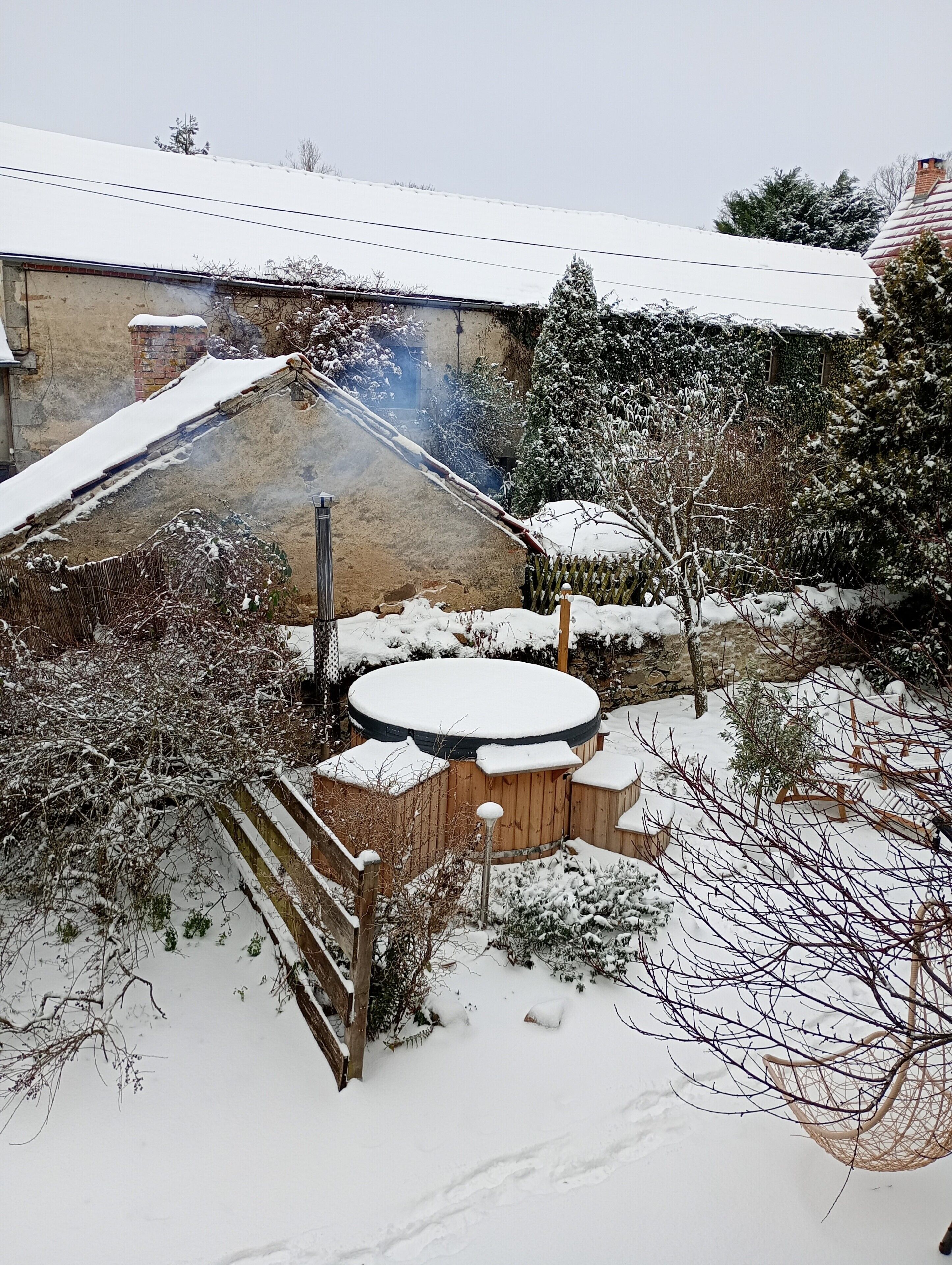 Un espacio exterior cubierto de nieve con un jacuzzi, valla de madera y un edificio con techo inclinado.