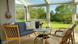 Een oranjerie met een bank, rieten stoelen, een glazen tafel en uitzicht op groen.