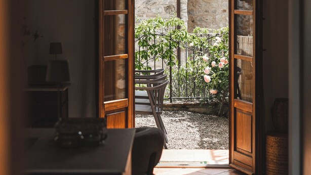 Una puerta abierta que da a un patio con una mesa y sillas, un pequeño jardín con plantas y un muro de piedra.