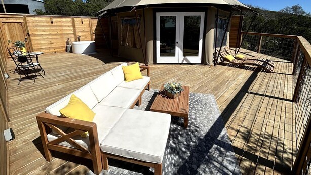 Ein überdachter Außenbereich mit Holzterrasse, einem weißen Sofa mit gelben Kissen und einem Holztisch.