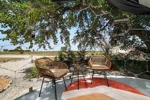 Un espace extérieur aménagé avec deux fauteuils en osier et une table, entouré d’arbres et sous un ciel dégagé.