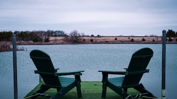 Two green Adirondack chairs on a dock over water with a grassy shoreline in the background.