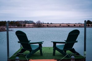 Two green Adirondack chairs on a dock over water with a grassy shoreline in the background.