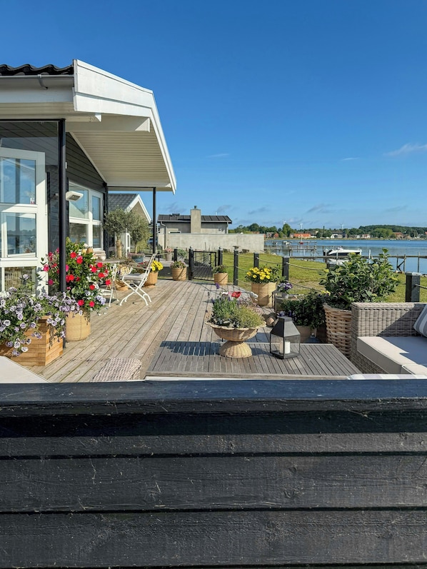 Une terrasse en bois avec des meubles d’extérieur, des plantes en pot et une vue sur un lac et des bâtiments en arrière-plan.