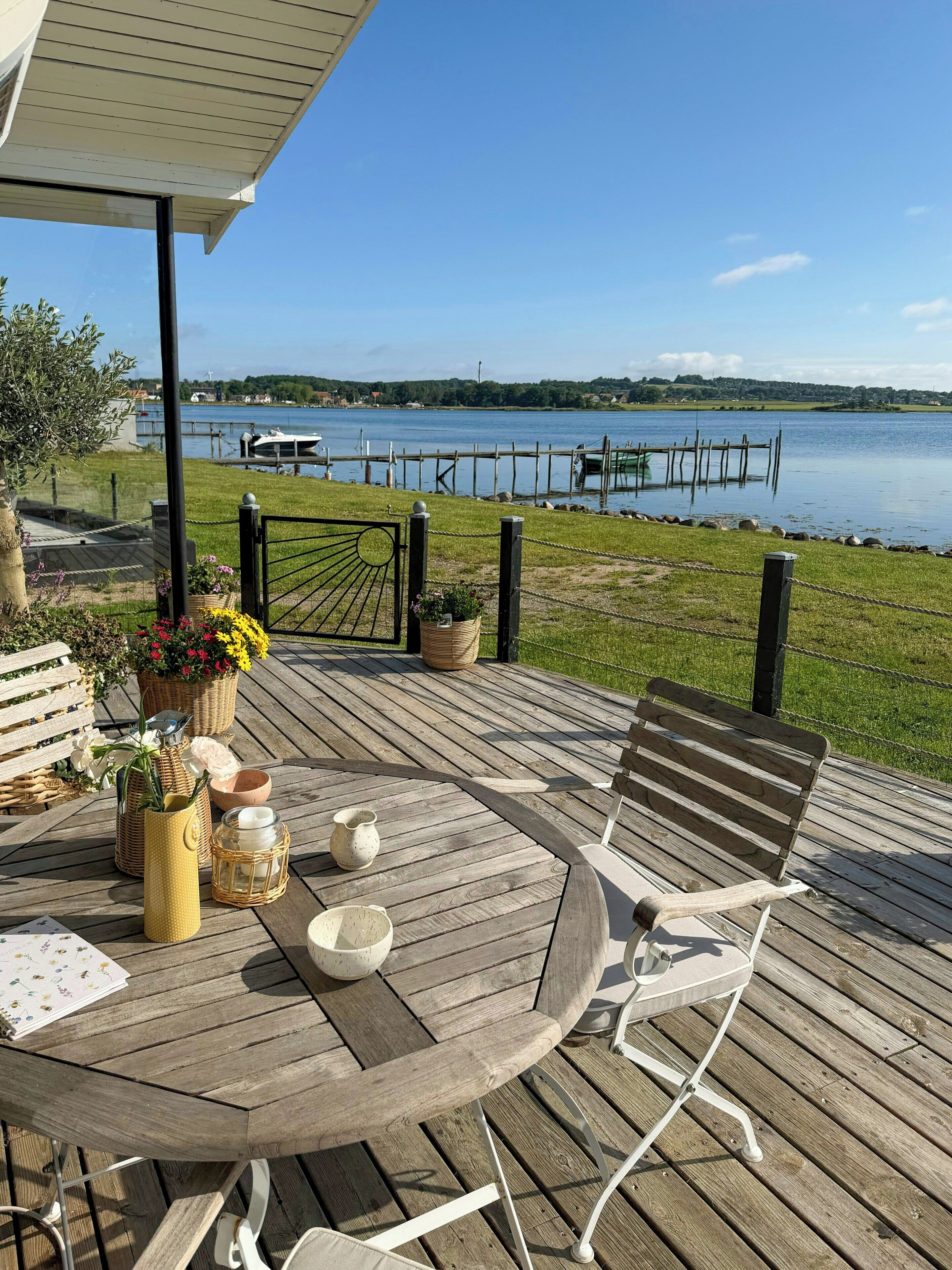 Una terraza de madera con una mesa y sillas, con vista a un lago con barcos y un muelle.