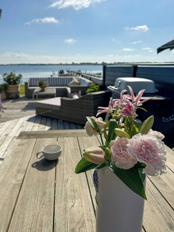A wooden deck with outdoor furniture, a white vase with flowers, and a view of the water.