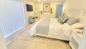 Une chambre à coucher comprenant un lit, des tables de chevet, un ventilateur de plafond, une télévision, une fenêtre avec des stores et une porte donnant sur une autre pièce.