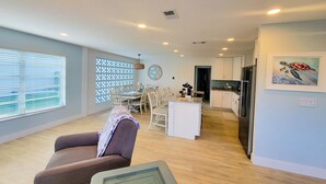 Un salon moderne avec un canapé, un coin repas comprenant une table et des chaises, une cuisine équipée de meubles et d’appareils électroménagers, et une grande fenêtre avec des stores.