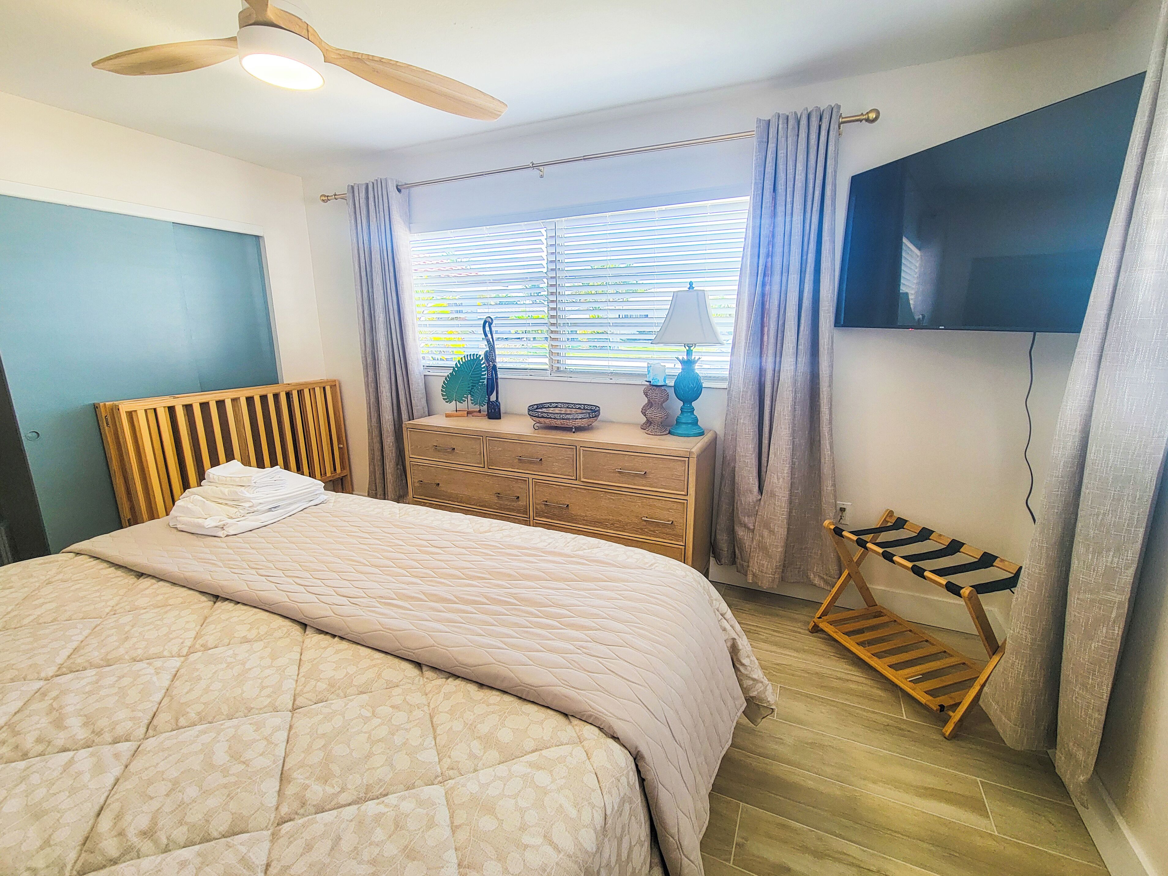 Une chambre à coucher avec un lit, une commode, un téléviseur et une fenêtre avec des rideaux.
