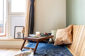 Ein gemütliches Zimmer mit einer hölzernen Bank, einem kleinen Tisch mit Buch und Kerze, einem gerahmten Bild und einem Fenster mit Vorhängen.