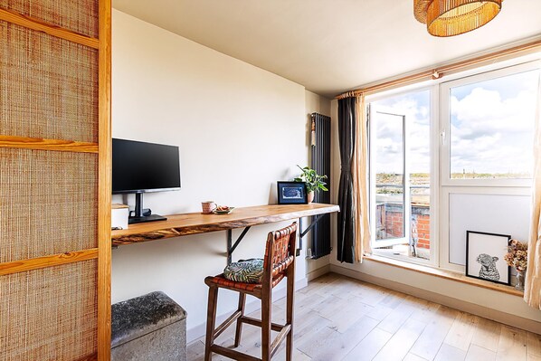Ein Zimmer mit einem Holztisch, einem Fernseher, einem Fenster mit Vorhängen, einer Topfpflanze und einem gerahmten Bild.