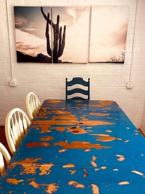A dining area with a blue table, white chairs, and a wall-mounted painting of cacti.