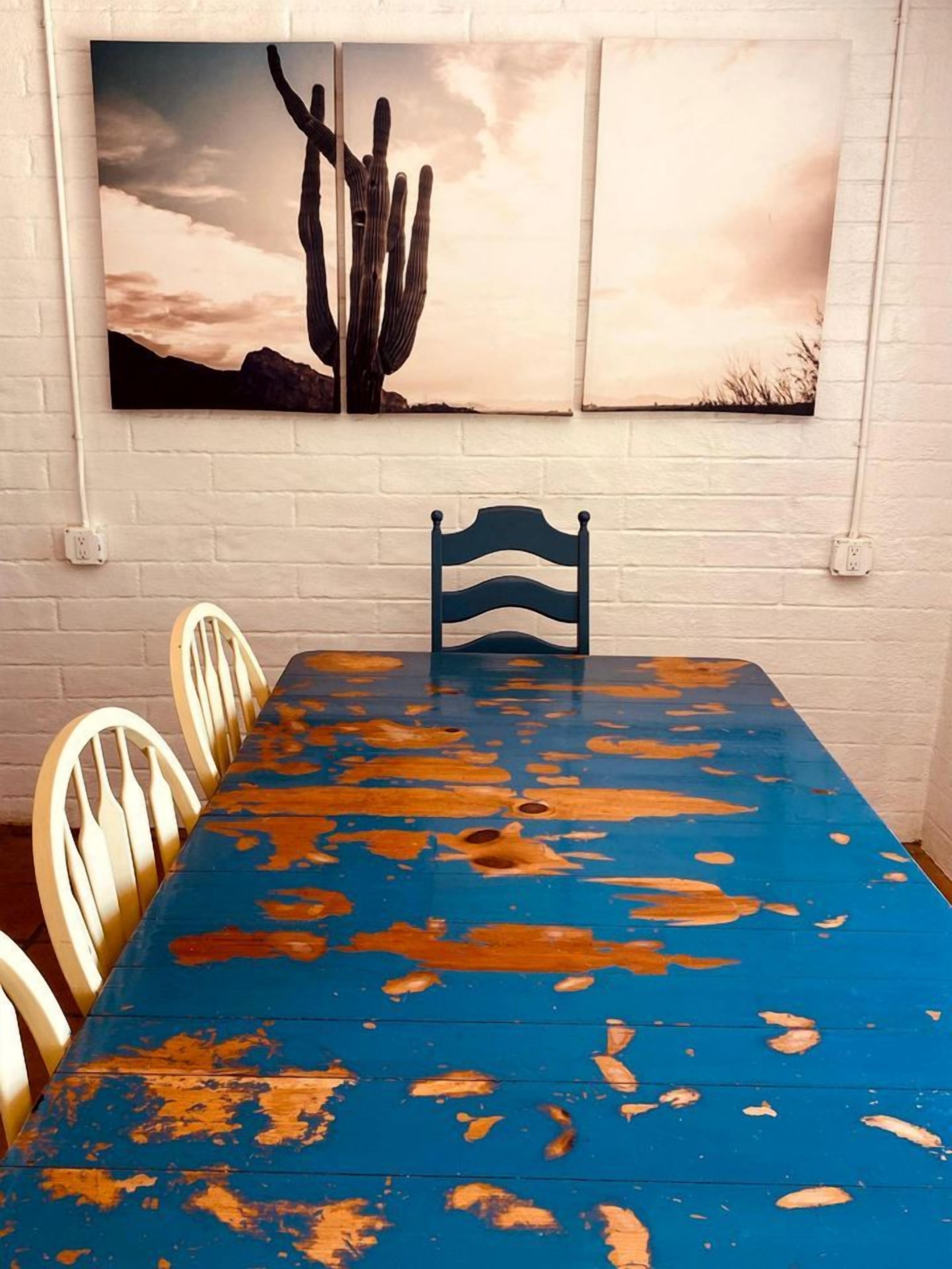 Uma área de refeições com mesa azul, cadeiras brancas e um quadro de cactos pendurado na parede.