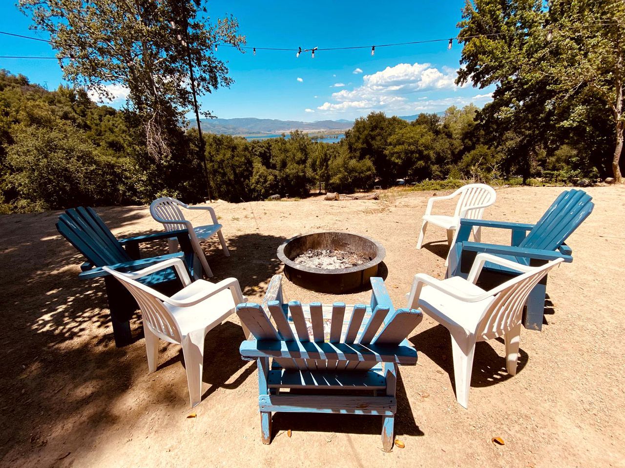 Een camping met vier Adirondack stoelen rond een vuurkuil, omgeven door bomen en een heldere lucht.