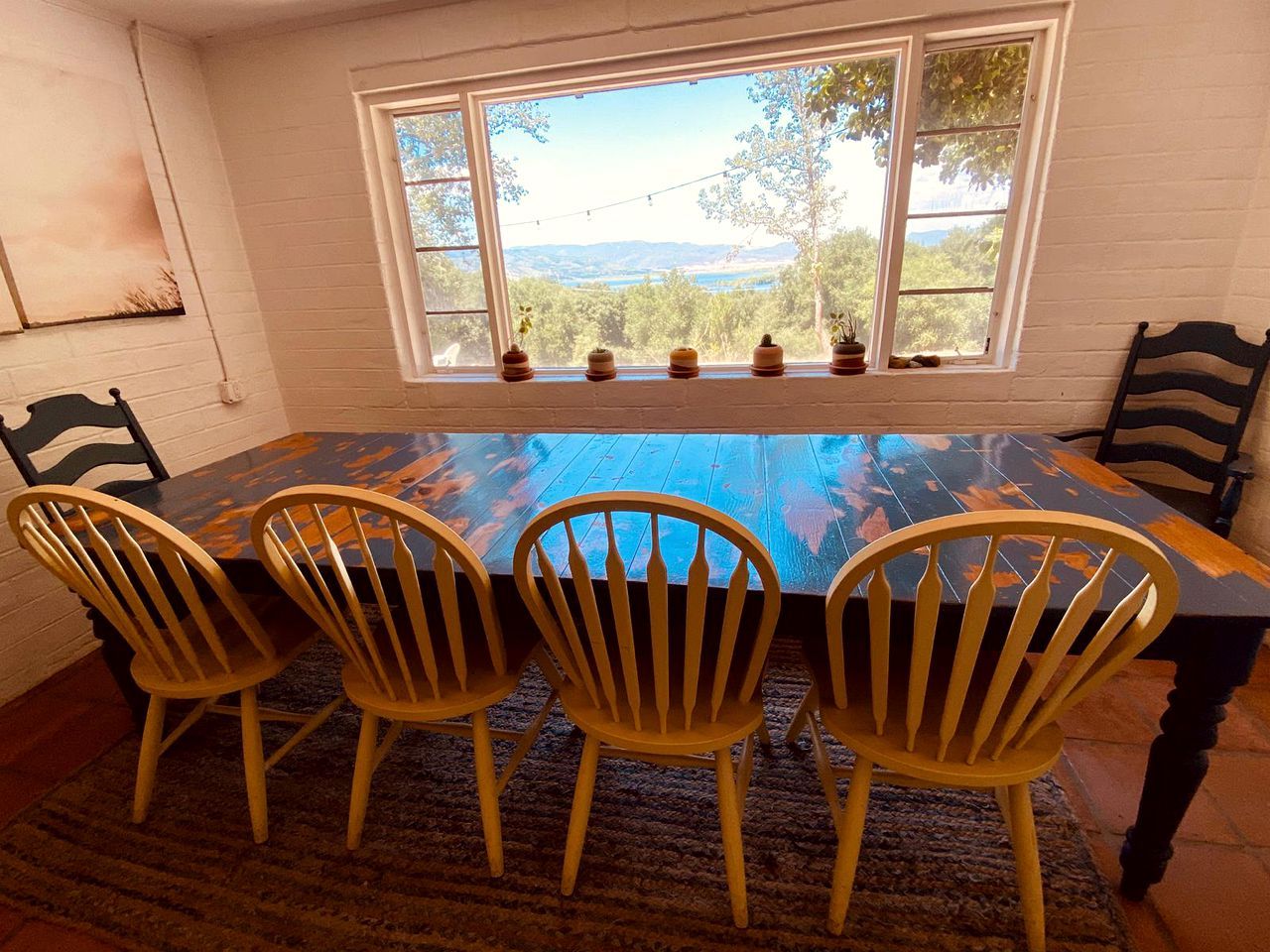 Une table à manger avec quatre chaises en bois, une fenêtre donnant sur un paysage arboré et montagneux, et un tapis sur le sol.