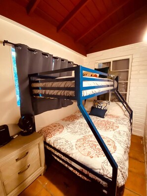Un llit superposat amb un estriat, una tauleta de nit amb una làmpada i una finestra amb cortines.
