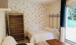Une petite pièce comprenant un lit, une étagère en bois, une fenêtre donnant sur un espace verdoyant et une porte avec un rideau.