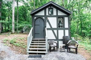Eine kleine, dunkel gefärbte Holzhütte mit einer weißen Tür und einem Fenster, umgeben von einem Wald. Vor der Tür führen Treppen hinauf und es stehen zwei Stühle bereit.