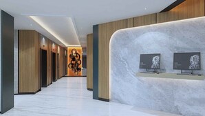 Un moderno vestíbulo de hotel con un mostrador de recepción de mármol, paneles de madera y un elemento decorativo de iluminación en el centro.
