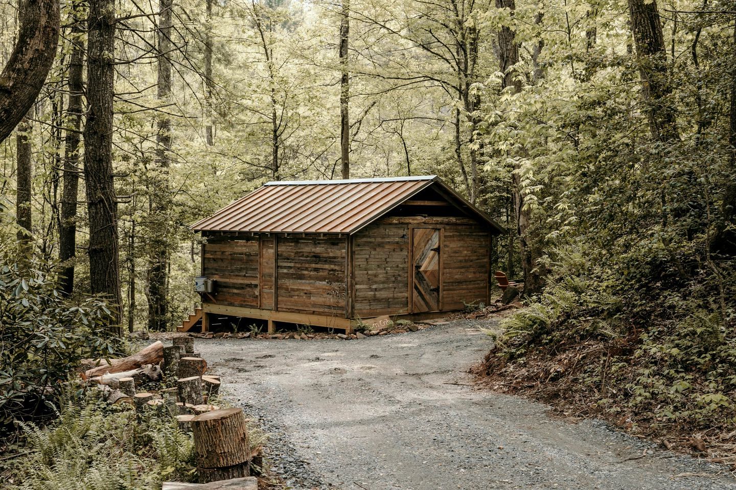 Une cabane en bois rustique, située dans une forêt, avec un chemin de gravier menant à l’entrée.