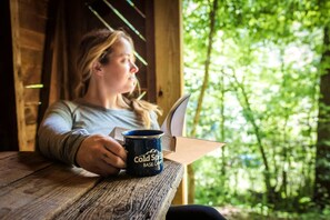 A person sitting at a wooden table with a book and a mug labeled 'Cold Spruce Base Camp.'