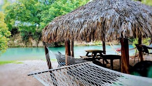 A hammock under a thatched roof structure by a calm body of water, with a picnic table and chairs nearby. - Fully-Equipped Riverside Glamping Safari Tent on the San Marcos River, Texas (Kingsbury)