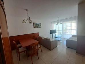 A living room with a sofa, a dining table with chairs, a TV, and a painting on the wall. - LSC Apartmenthotel (Siófok)