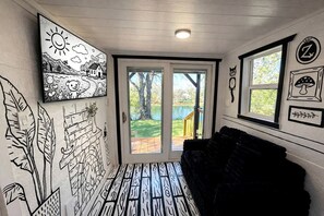 Quarto com sofá preto, porta que dá para o exterior e arte nas paredes, incluindo um sol, um urso e um cogumelo.