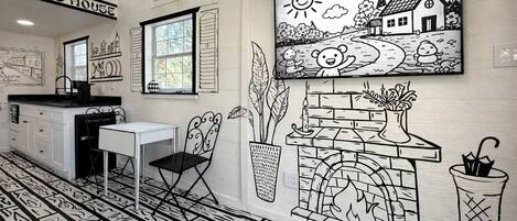 A room with a fireplace, a dining table, a chair, a window, and a wall with a black and white mural.