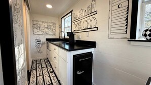 A kitchen with a black countertop, white cabinets, and a refrigerator. There are black and white wall decorations, including a welcome sign and a character illustration.