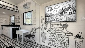A room with a fireplace, a dining table, a chair, a window, and a wall with a black and white mural.