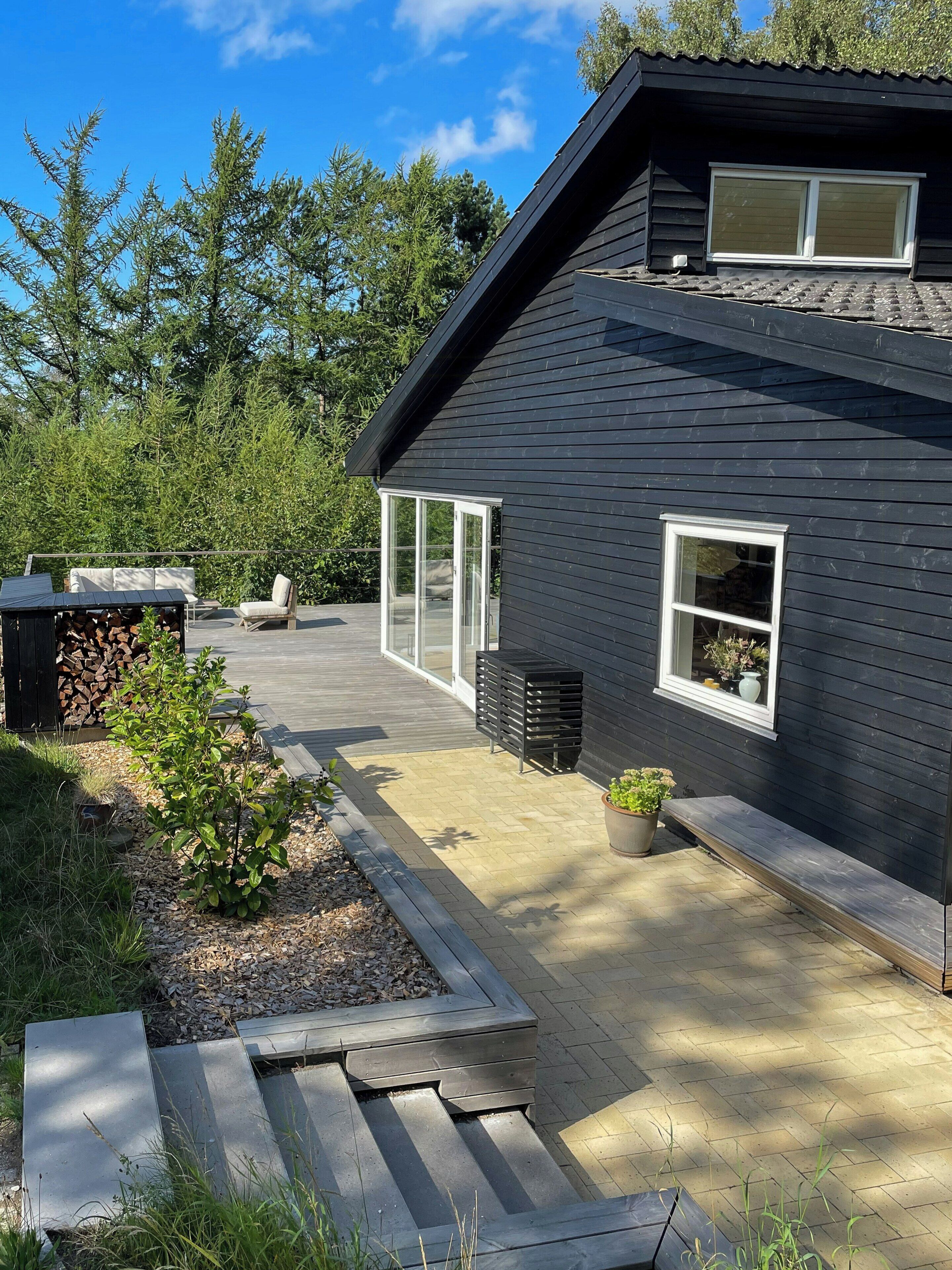A modern house with a patio, a stack of firewood, a bench, and a small garden area.