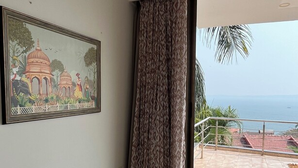 A room with a large window, a framed painting, and a view of a building and palm trees.