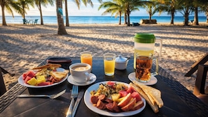 Desayuno en la playa con una mesa, platos de comida, bebidas y vistas al océano y palmeras.