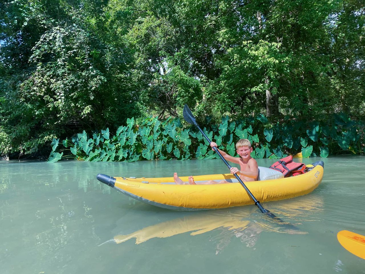A person in a yellow inflatable raft on a calm river surrounded by dense green foliage.