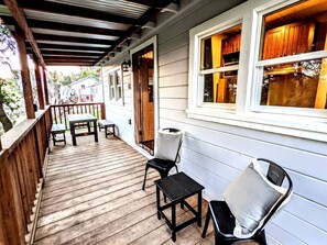 A covered porch with wooden flooring, two chairs, a small table, and a window with wooden shutters.