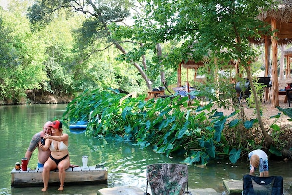 A couple sitting on a dock by a river, surrounded by lush greenery and a thatched-roof structure.