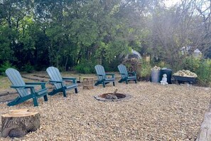 A fire pit surrounded by four Adirondack chairs in a wooded outdoor area.