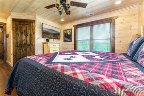 A bedroom with a large bed, wooden walls, a ceiling fan, a window with blinds, and a door. - Outstanding Cabin with Game Room, Jacuzzi and Cinema Room in Tennessee (Sevierville)