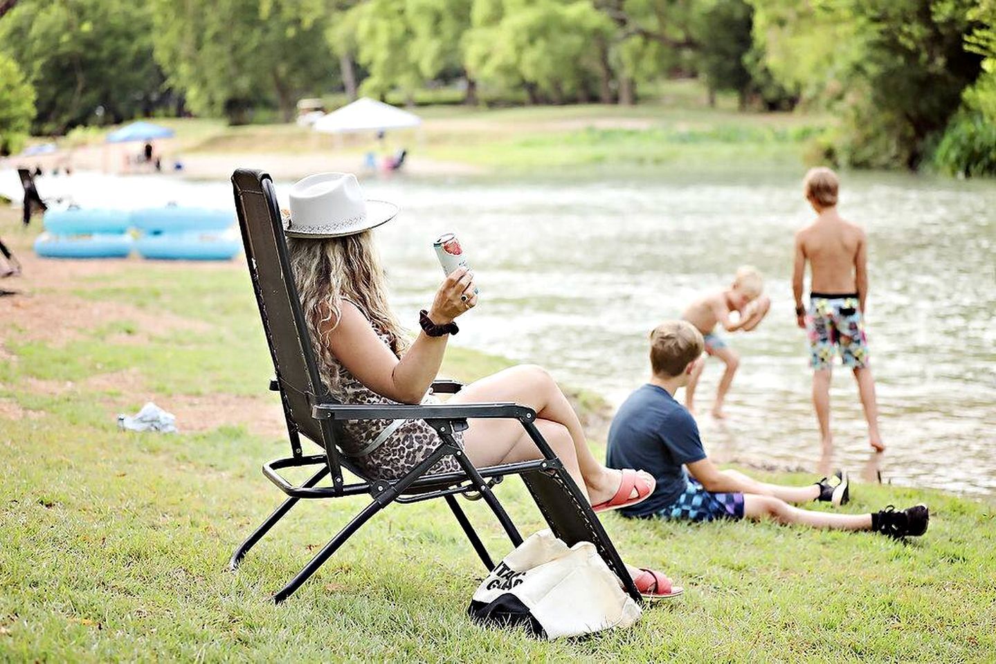 A person sitting on a lawn chair by a lake, another person sitting on the grass, and a third person standing in the water.