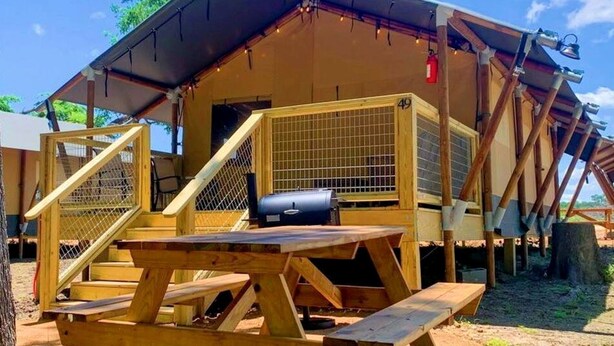 A wooden structure with a picnic table and grill, a fenced area, and a building with a number '49' visible. - Texas Glamping at its Finest in Safari Tents on the San Marcos River (Kingsbury)