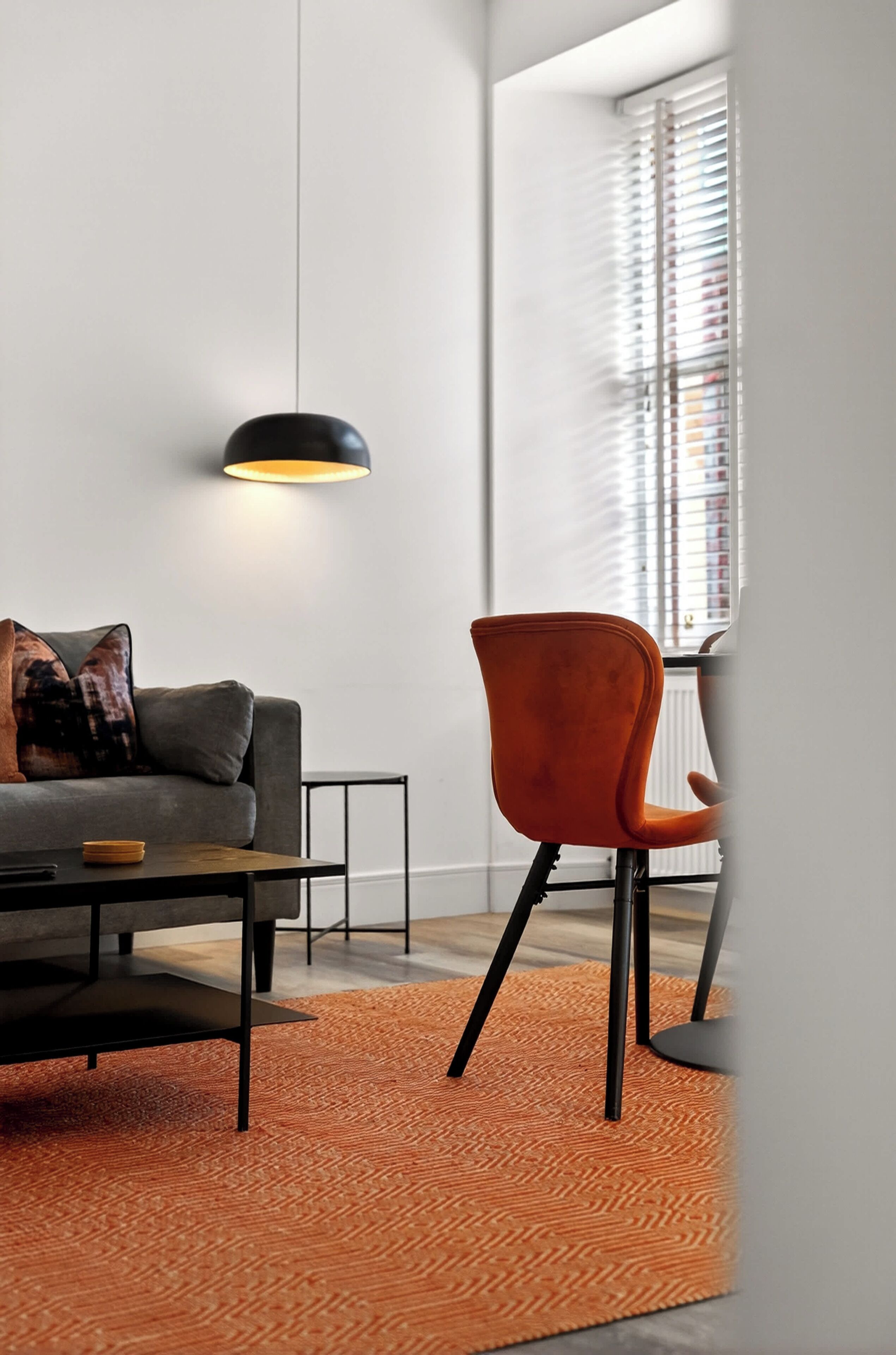 A modern living room with a grey sofa, orange chair, black pendant light, and a patterned orange rug.