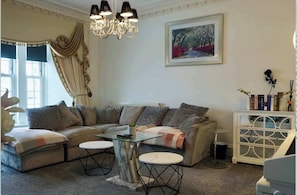 A living room with a sectional sofa, a glass coffee table, a chandelier, a framed painting, and a white cabinet with books.