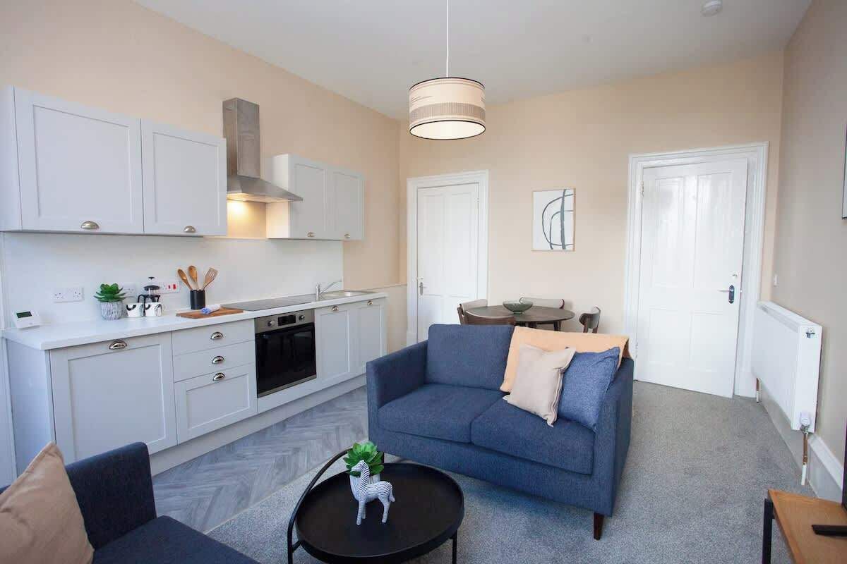 A modern apartment with a kitchenette, a blue sofa, a small dining table, and a pendant light.